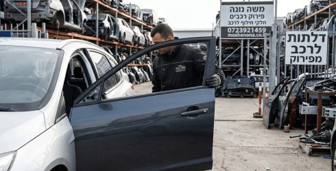 מבט על מגרש פירוק רכבים של משה נונה ובו עובד מרכיב או בודק דלת אפורה כהה של מכונית כסופה בחזית התמונה כאשר ברקע נראים מדפי ענק עמוסים בשלדות רכבים וחלקי חילוף שלטים בעברית עם פרטי קשר והכיתוב דלתות לרכב מפירוק לצד שורת דלתות מפורקות המונחות על הקרקע